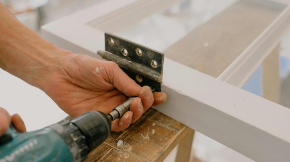 Crop unrecognizable worker drilling metal hinge on white window frame during house renovation